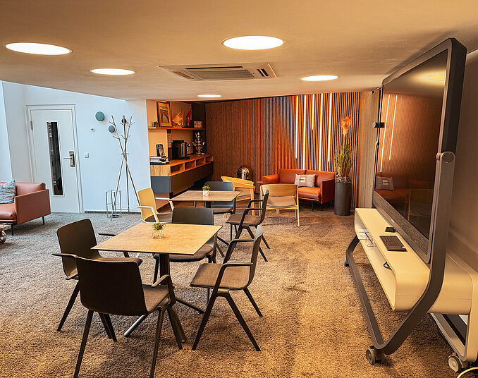 Moderner Tagungsraum mit flexiblen Tischgruppen, großem Präsentationsbildschirm, Sitzlounge und beleuchteter Holzwand