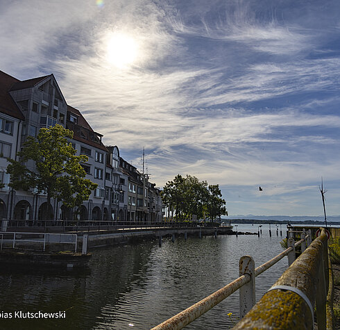 Sonnenlicht über Hafenpromenade mit Altbauten und ruhigem Wasser in Friedrichshafen.