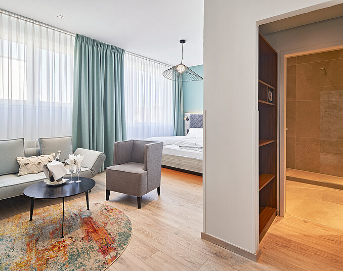 Ein helles und modernes Zimmer mit einem gemütlichen Sitzbereich, bestehend aus einem Sofa und einem Sessel. Ein runder Tisch steht in der Mitte. Im Hintergrund ist ein Bett sichtbar, und eine Dusche ist durch eine Tür abgetrennt. Große Fenster sorgen für viel Licht.