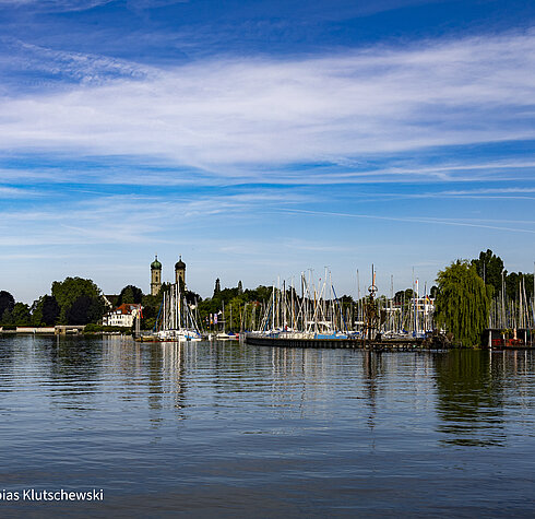 Blick auf den Bodensee mit Segelbooten im Hafen und den markanten Zwillingstürmen der Basilika St. Georg in Wasserburg.