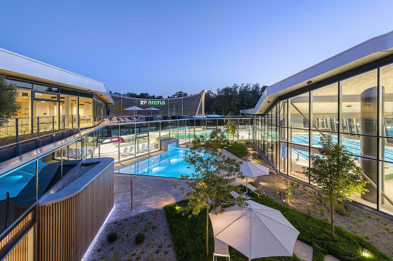 Eine moderne Freizeitanlage mit mehreren Schwimmbecken, umgeben von grünen Gartenbereichen und Bäumen. Die Architektur ist zeitgenössisch, mit viel Glas und Licht. Die Aufnahme zeigt die Anlage bei Dämmerung, was eine entspannende Atmosphäre vermittelt.