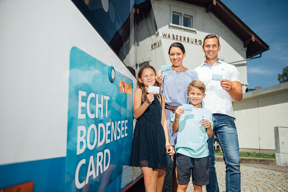 Eine Familie steht vor einem Bus und hält Bodensee-Karten in der Hand. Die Eltern und ihre zwei Kinder lächeln in die Kamera, während im Hintergrund ein Gebäude mit dem Schriftzug „Wasserburg“ zu sehen ist.