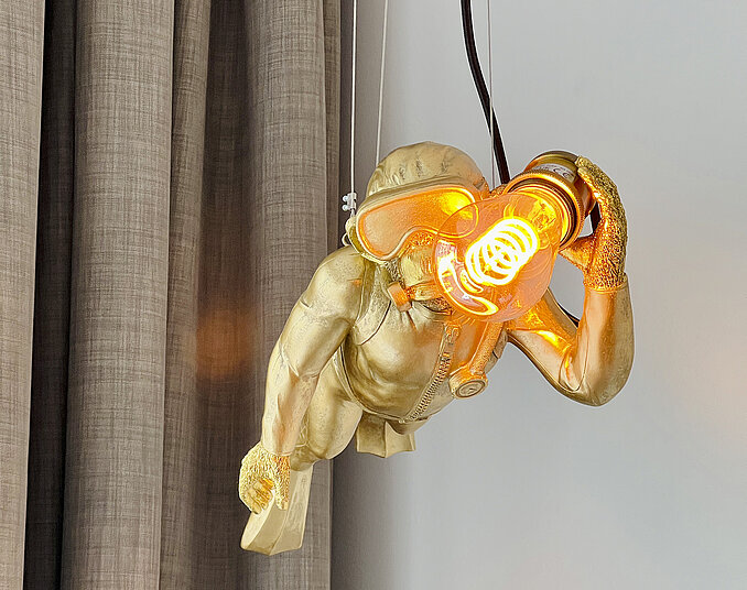 Ausgefallene Hängelampe in Form eines goldenen Tauchers mit leuchtender Taucherbrille vor einem Vorhang.
