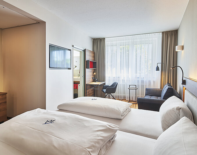 Das Bild zeigt ein modernes Hotelzimmer mit einem Doppelbett, einem Fenster mit Vorhängen, einem gemütlichen Sessel und einem kleinen Tisch. An einer Wand befindet sich ein Flachbildfernseher und ein Ablagetisch mit einem Wasserkessel und Snacks. Die Farben sind hell und freundlich.