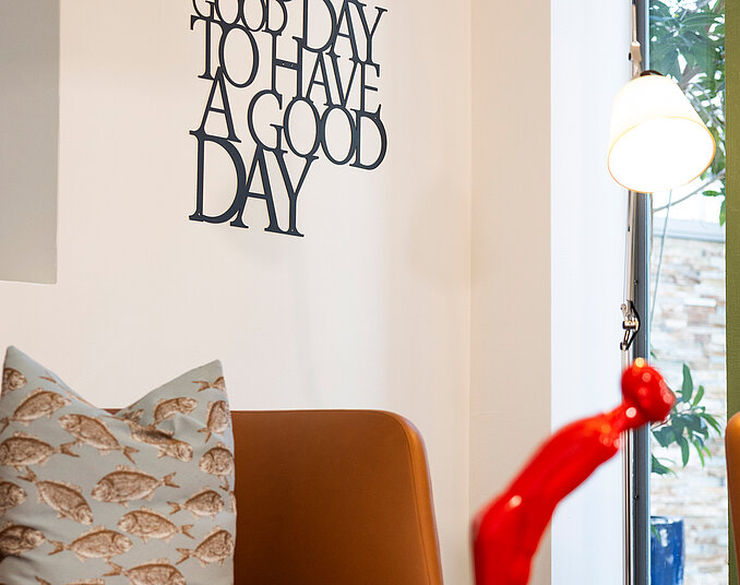 In einem modernen Raum steht ein brauner Sessel mit einem Kissen, das Fischmuster zeigt. An der Wand hängt eine schwarze Schrift mit dem Text "IT'S A GOOD DAY TO HAVE A GOOD DAY". In der Nähe befindet sich eine rote Skulptur und ein Tisch mit einer Lampe.