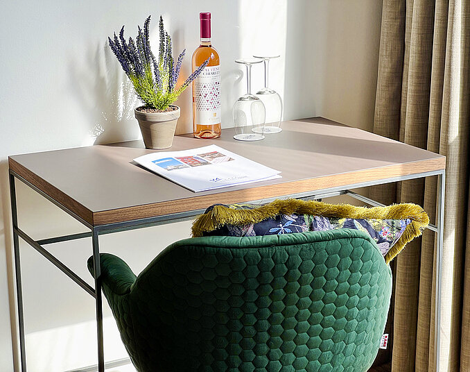 Ein modernes Schreibtisch-Setup mit einem hellen Holztablett, darauf steht eine Flasche Roséwein, zwei Gläser und eine kleine Blume in einem Topf. Daneben liegt eine Zeitschrift und ein grüner Stuhl mit einem bunten Kissen ist sichtbar. Vor dem Tisch sind Vorhänge zu sehen.