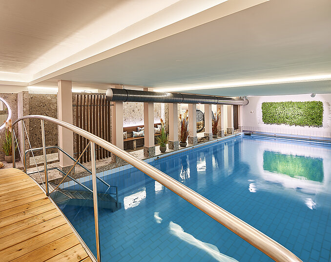 Ein moderner Innenpool mit klarem, blauem Wasser, umgeben von Pflanzen und natürlichen Materialien. Eine Holzterrasse und eine Stiege führen ins Wasser. Die Beleuchtung ist sanft und schafft eine entspannende Atmosphäre.