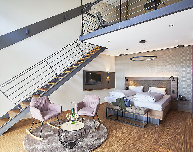 Ein modernes Hotelzimmer mit einer Schlafgelegenheit, einem Fernseher und einer Sitzecke. Zwei Sessel stehen an einem kleinen Tisch, daneben ein Teppich. Eine Treppe führt in einen oberen Bereich des Raumes. Die Wände sind in neutralen Farben gehalten.