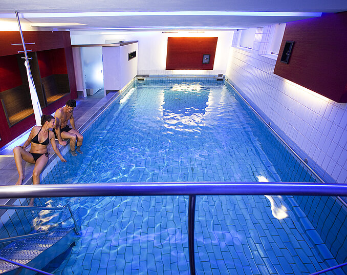 Zwei Personen sitzen am Rand eines Innenpools. Das Wasser ist klar und reflektiert das Licht von bunten Wänden. Die Umgebung wirkt modern und einladend, mit Fliesen und einer Treppe zum Wasser.