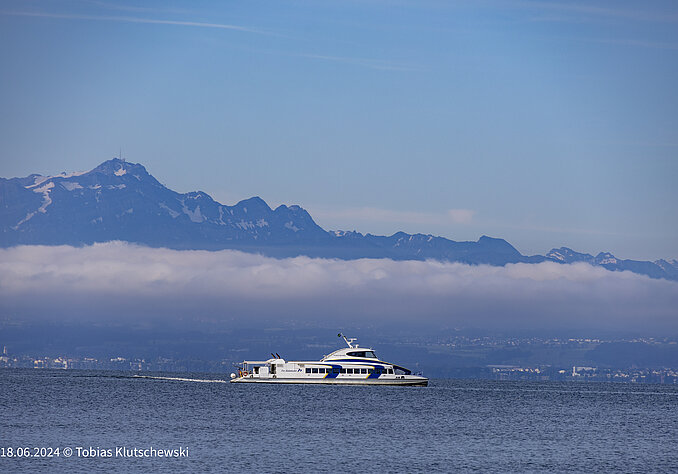 Ein Passagierschiff fährt auf einem ruhigen See, während im Hintergrund die majestätischen Berge und eine Wolkenbank zu sehen sind. Der Himmel ist klar und hellblau.