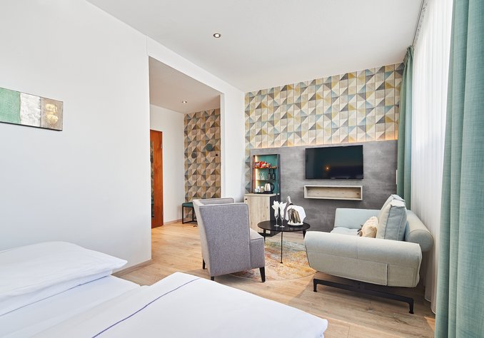 Ein modernes Zimmer mit einem Doppelbett, einem gemütlichen Wohnzimmerbereich, einem Fernseher an der Wand und einer dekorativen Wand mit geometrischem Muster. Große Fenster lassen viel Licht hinein, und es gibt einen stilvollen Sessel sowie einen Tisch im Raum.