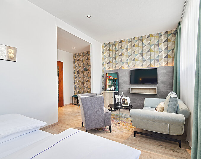 Ein modernes Zimmer mit einem Doppelbett, einem gemütlichen Wohnzimmerbereich, einem Fernseher an der Wand und einer dekorativen Wand mit geometrischem Muster. Große Fenster lassen viel Licht hinein, und es gibt einen stilvollen Sessel sowie einen Tisch im Raum.