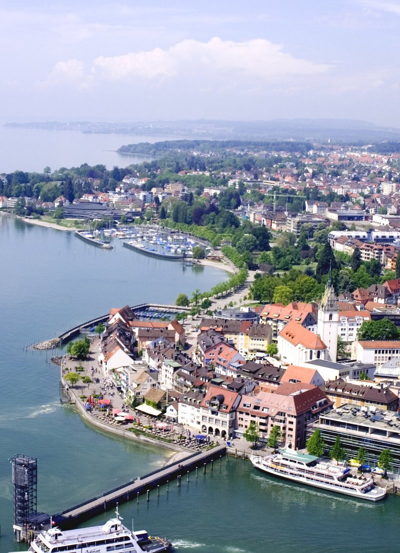 Luftaufnahme einer Küstenstadt am Bodensee, mit Aussicht auf Wohngebäude, Parks, Marina und Uferpromenade. Der Himmel ist blau mit einigen Wolken, und das Wasser reflektiert das Licht.