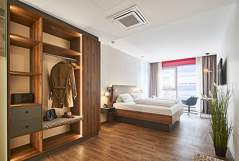 Modernes Hotelzimmer mit offenem Kleiderschrank, gemütlichem Doppelbett und großem Fenster mit Stadtblick.