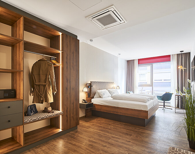 Modernes Hotelzimmer mit offenem Kleiderschrank, gemütlichem Doppelbett und großem Fenster mit Stadtblick.