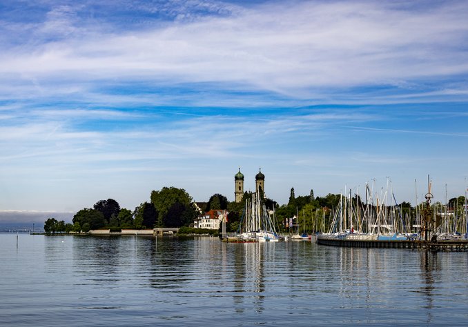 Blick auf einen ruhigen See mit einem Hafen, in dem mehrere Segelboote liegen. Im Hintergrund sind grüne Bäume und zwei Türme eines Gebäudes sichtbar, unter einem blauen Himmel mit wenigen Wolken.