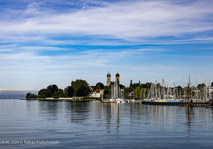 Blick auf einen ruhigen See mit einem Hafen, in dem mehrere Segelboote liegen. Im Hintergrund sind grüne Bäume und zwei Türme eines Gebäudes sichtbar, unter einem blauen Himmel mit wenigen Wolken.