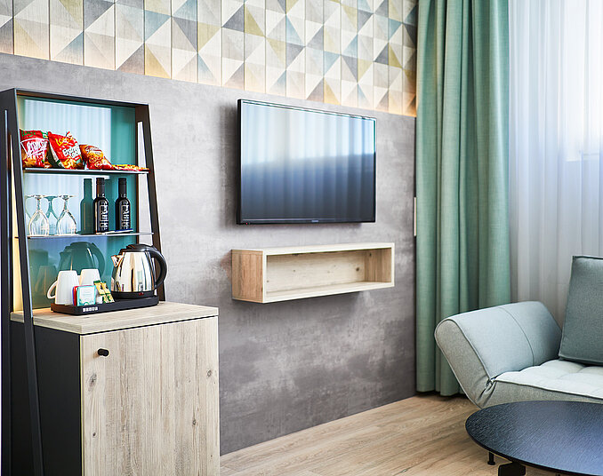 Ein modernes Zimmer mit einem Fernseher an der Wand, einem eleganten Sofa und einem Tisch. In einer Ecke steht ein kleiner Kühlschrank mit Snacks und Getränken, daneben eine Teekanne und Tassen. Der Raum hat eine stilvolle Wandgestaltung und große Fenster mit Vorhängen.