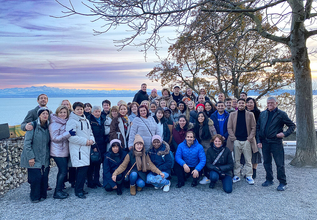 Eine große Gruppe von Menschen posiert fröhlich vor der malerischen Kulisse des Bodensees bei Sonnenuntergang.
