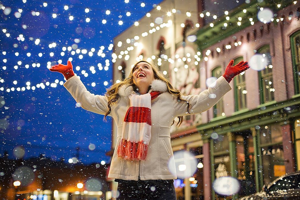 Eine fröhliche Frau steht in einer festlich beleuchteten Straße und genießt den Schnee, der um sie herum fällt. Sie trägt einen warmen Mantel und einen gestreiften Schal und breitet lächelnd die Arme aus. Im Hintergrund sind Lichter und historische Gebäude sichtbar.