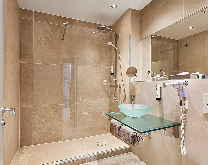 Moderne Badezimmeransicht mit einer Dusche, die durch Glasabtrennungen vom restlichen Raum getrennt ist. Eine gläserne Waschschüssel steht auf einer transparenten Ablage. Fliesen in neutralen Farbtönen dominieren die Wände und den Boden. Ein Föhn und andere Toilettenartikel sind ebenfalls sichtbar.