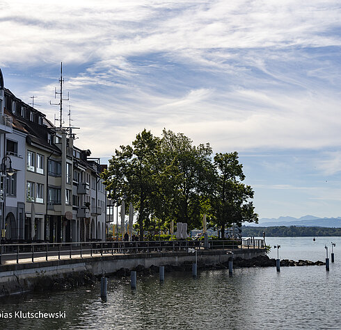 Uferpromenade am Bodensee mit Blick auf das Wasser und die Alpen im Hintergrund an einem sonnigen Tag.