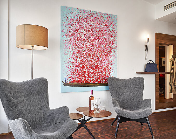 Zwei graue Sessel stehen neben einem kleinen Tisch, auf dem eine Flasche Rosé-Wein und zwei Gläser stehen. Im Hintergrund hängt ein großes, farbenfrohes Gemälde mit roten Blüten auf blauem Grund. Eine Stehlampe und ein Kleiderständer sind ebenfalls im Raum sichtbar.