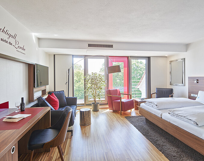 Das Bild zeigt ein modernes Hotelzimmer mit einem Doppelbett, einem Schreibtisch, einem Sessel und großen Fenstern, die natürliches Licht hereinlassen. An der Wand befindet sich ein Zitat. Der Stil ist minimalistisch und freundlich mit warmen Holztönen.