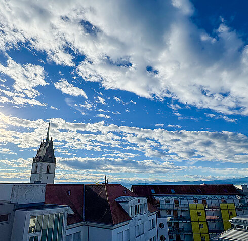 Blick über die Dächer von Friedrichshafen mit der Schlosskirche und dem Bodensee im Hintergrund unter einem blauen Himmel.
