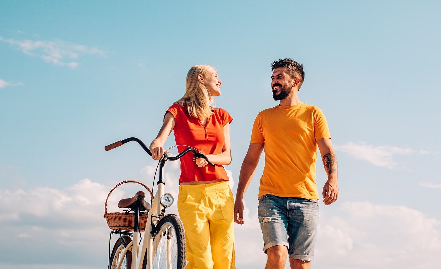 Fröhliches Paar spaziert bei sonnigem Wetter, die Frau schiebt ein Fahrrad mit Korb, beide lachen sich an.
