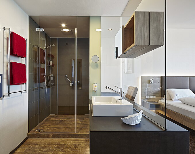 Modernes Hotelbadezimmer mit offener Glasdusche, stilvollem Waschbecken und Blick ins Schlafzimmer.