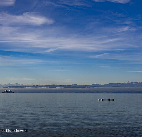 Blick über den Bodensee mit einer Fähre in der Ferne und den Alpen im Hintergrund unter einem leicht bewölkten Himmel.