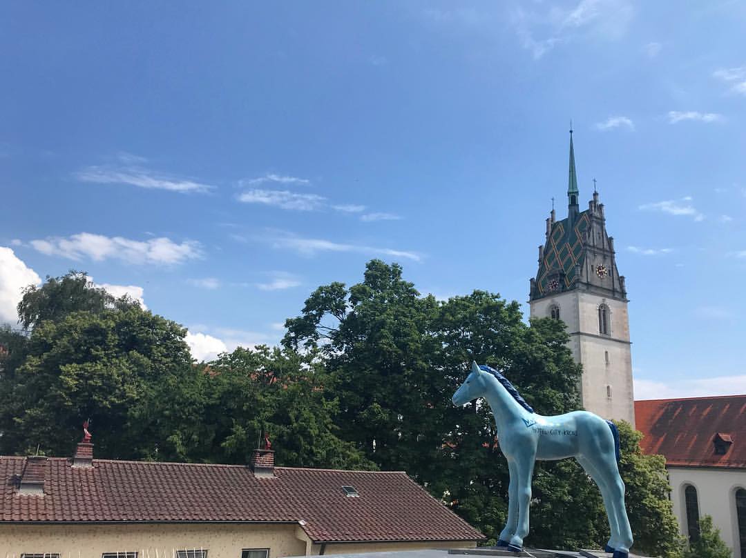 Blaues Pferdekunstwerk auf einem Dach vor grünen Bäumen und einem historischen Kirchturm unter blauem Himmel.