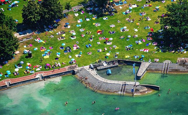 Eine luftige Aufnahme eines Strandes mit vielen Menschen auf der Wiese. Bunte Handtücher und Sonnenschirme sind verteilt, während mehrere Badegäste im klaren Wasser schwimmen. Im Hintergrund sind Bäume zu sehen, die dem Ort Schatten spenden.