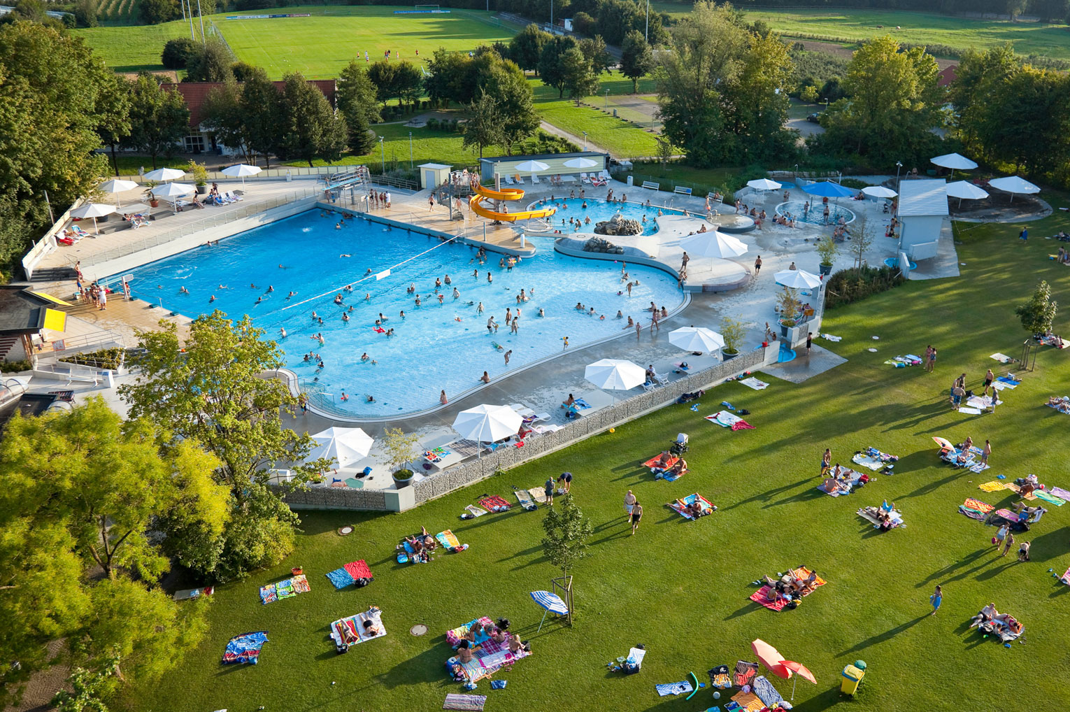 Eine Luftaufnahme eines Schwimmbades mit einem großen Becken, in dem viele Menschen schwimmen und spielen. Rund um das Becken stehen Sonnenschirme und Liegen auf einer grünen Wiese. Bäume und ein üppiges Gelände umgeben den Bereich.