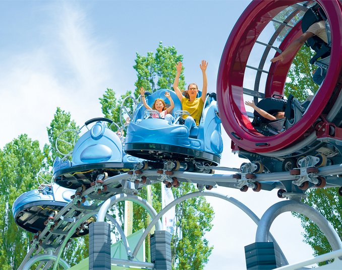 Zwei Kinder und ein Erwachsener fahren fröhlich in einem blauen Fahrgeschäft auf einer Achterbahn. Sie heben die Hände in die Luft und genießen die Fahrt in einer Freizeitpark-Umgebung mit Bäumen im Hintergrund.