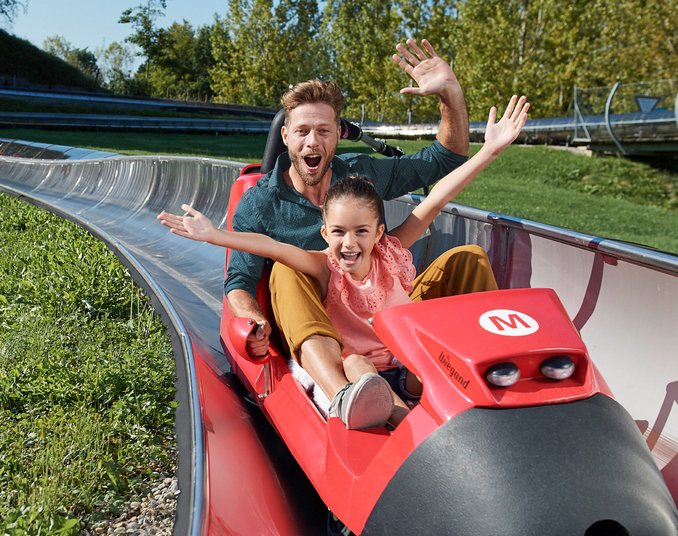Ein Mann und ein Mädchen sitzen glücklich in einer roten Sommerrodelbahn. Beide haben die Arme in die Luft gehoben und lächeln, während sie rasch den Hang hinunterfahren. Im Hintergrund sind grüne Bäume und eine Rodelbahn zu sehen.