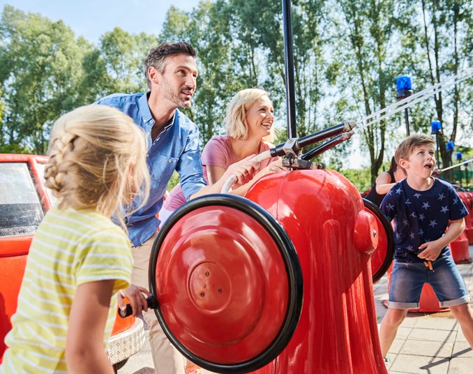 Eine Familie mit zwei Erwachsenen und zwei Kindern spielt an einem Wasserspielplatz. Sie bedienen rote Wasserpumpen, während Wasser spritzt. Im Hintergrund sind Bäume und Spielgeräte zu sehen. Sonne scheint und die Stimmung ist fröhlich.