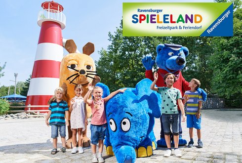 Eine Gruppe von Kindern steht vor einem hohen Leuchtturm und posiert mit zwei großen, bunten Maskottchen: einem blauen Elefanten und einem braunen Tier. Im Hintergrund ist ein Schild mit der Aufschrift „Ravensburger SPIELELAND - Freizeitpark & Feriendorf“ sichtbar.