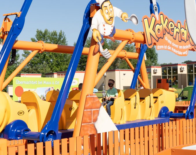 Ein bunter Freizeitpark-Attraktion namens "Kakerlaken" ist zu sehen. Sie besteht aus einer gelben, insektähnlichen Bahn mit einem fröhlichen Koch in einem weißen Outfit, der darauf steht. Die Bahn ist von einer orangefarbenen Umzäunung umgeben und in einem sonnigen Außenbereich platziert.